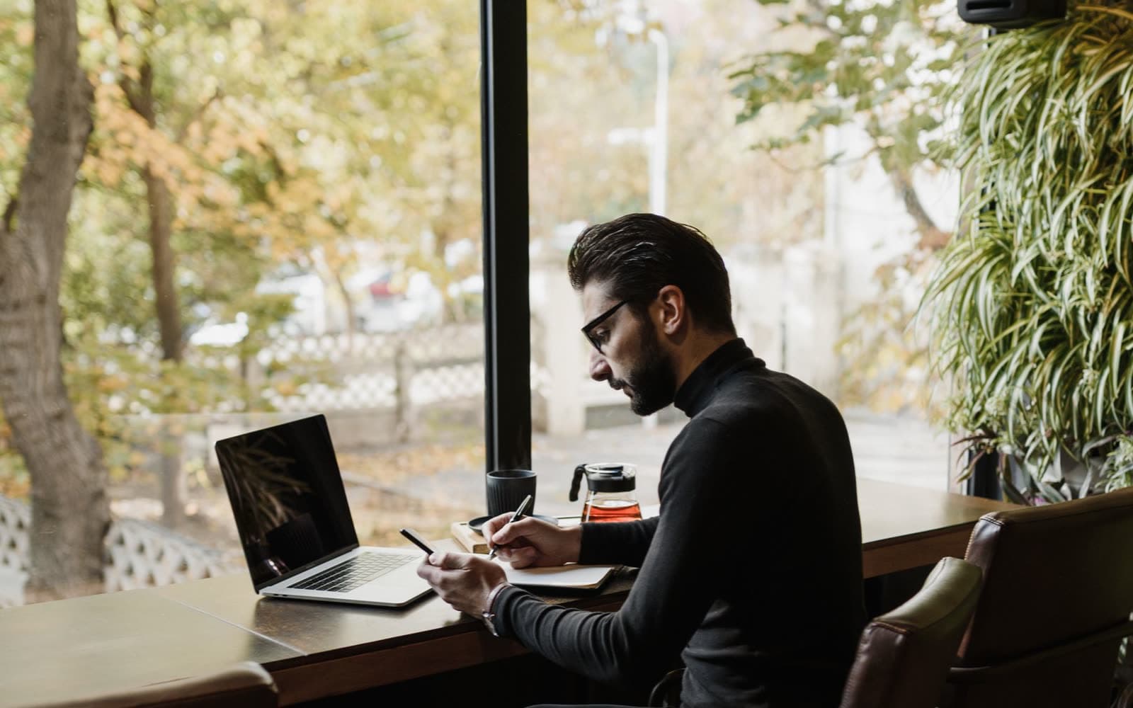 A person writing in a notebook beside an open laptop at a desk