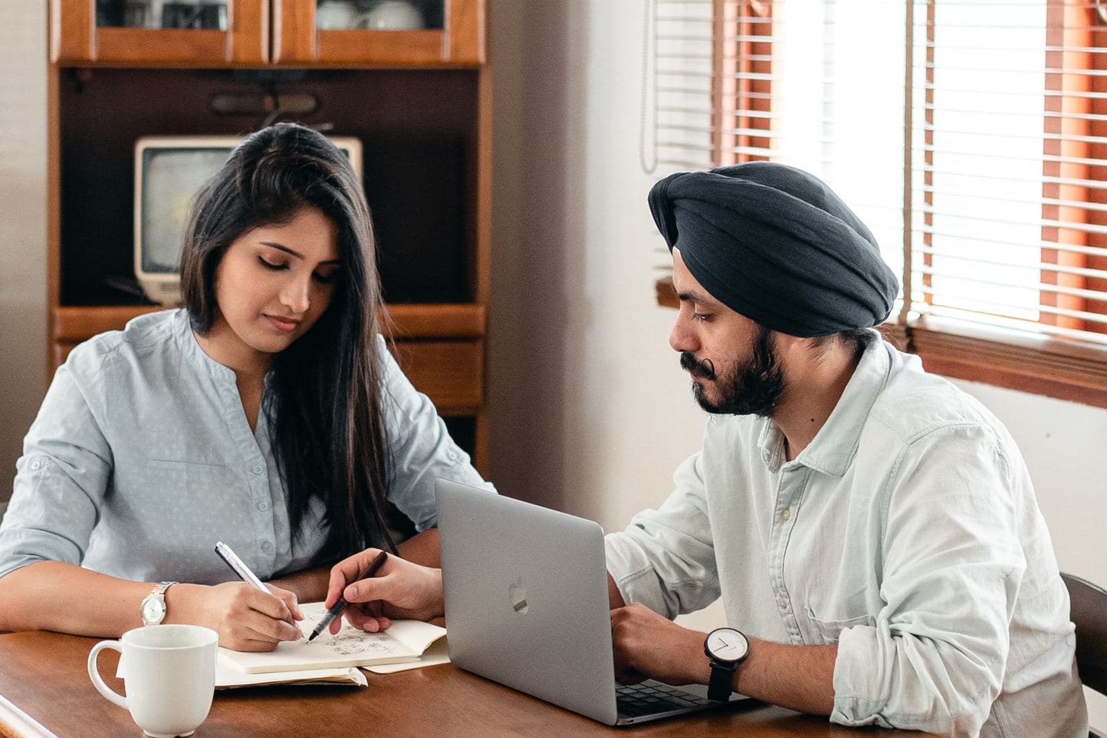 Two people at a table, one writing in a notebook and one using a laptop